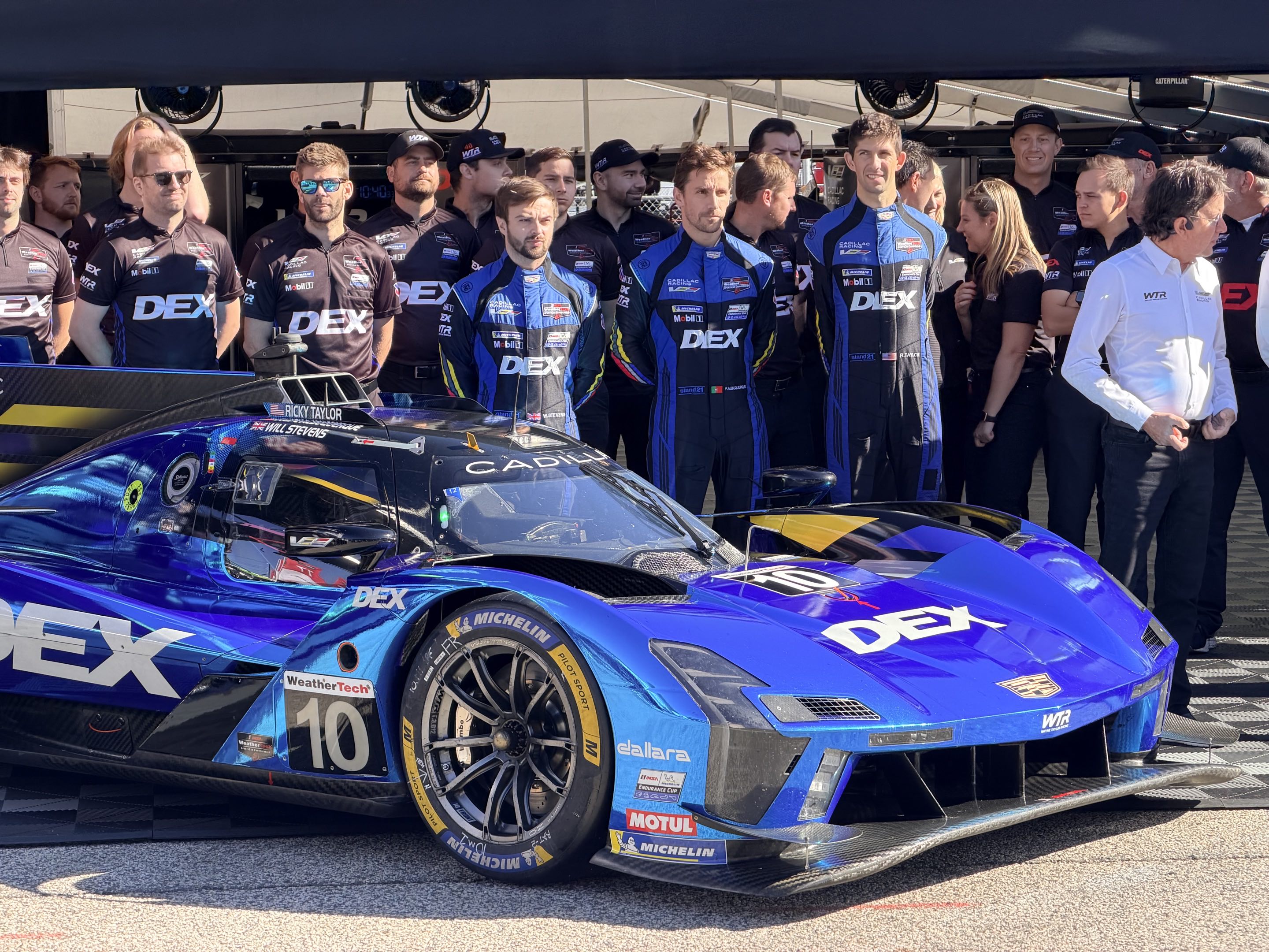 All the personnel of a Cadillac LMDh race team lined up behind the car facing left to right